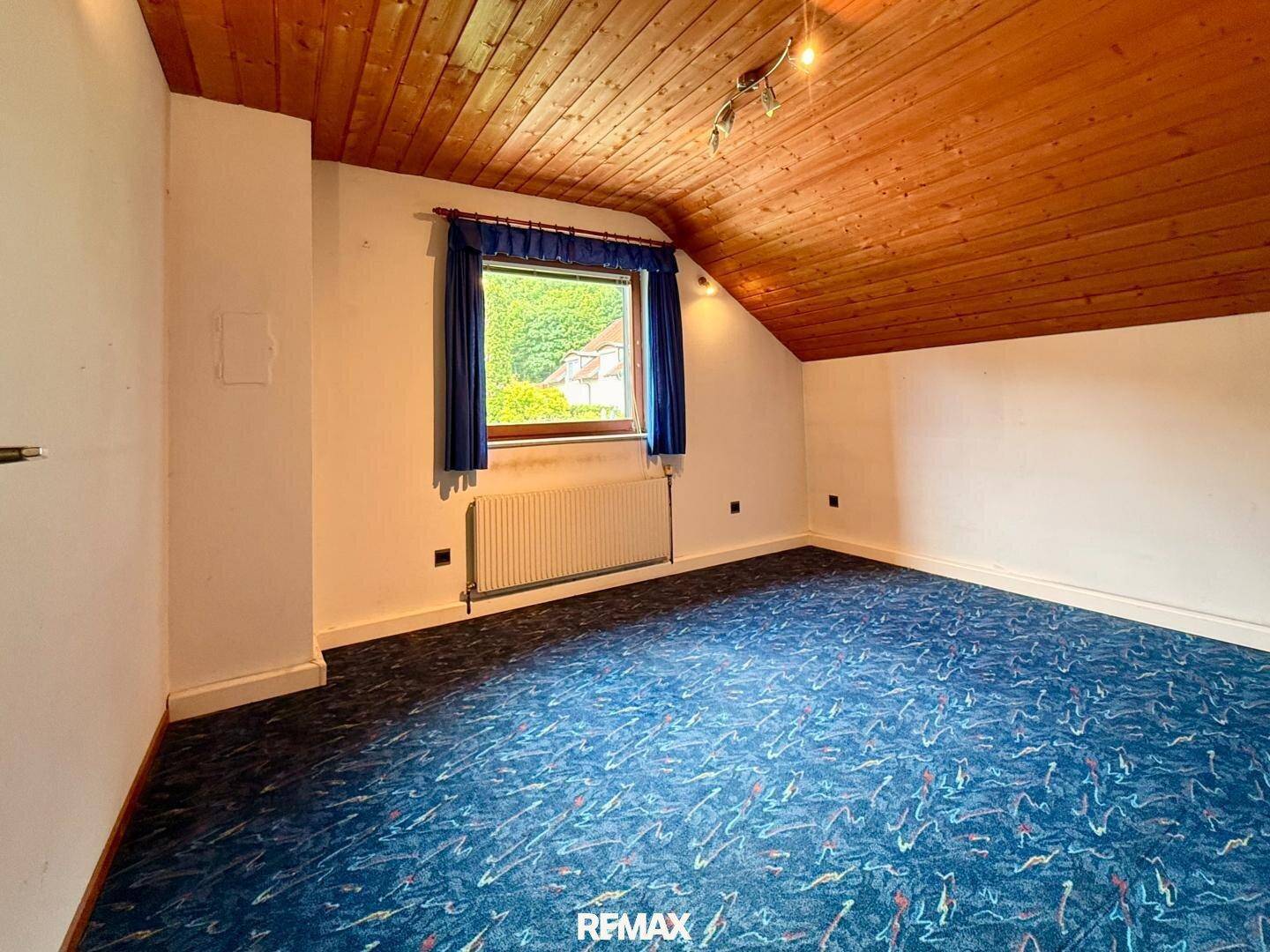Schlafzimmer im Dachgeschoss mit blauem Teppichboden, Holzdecke und Fenster mit Ausblick.