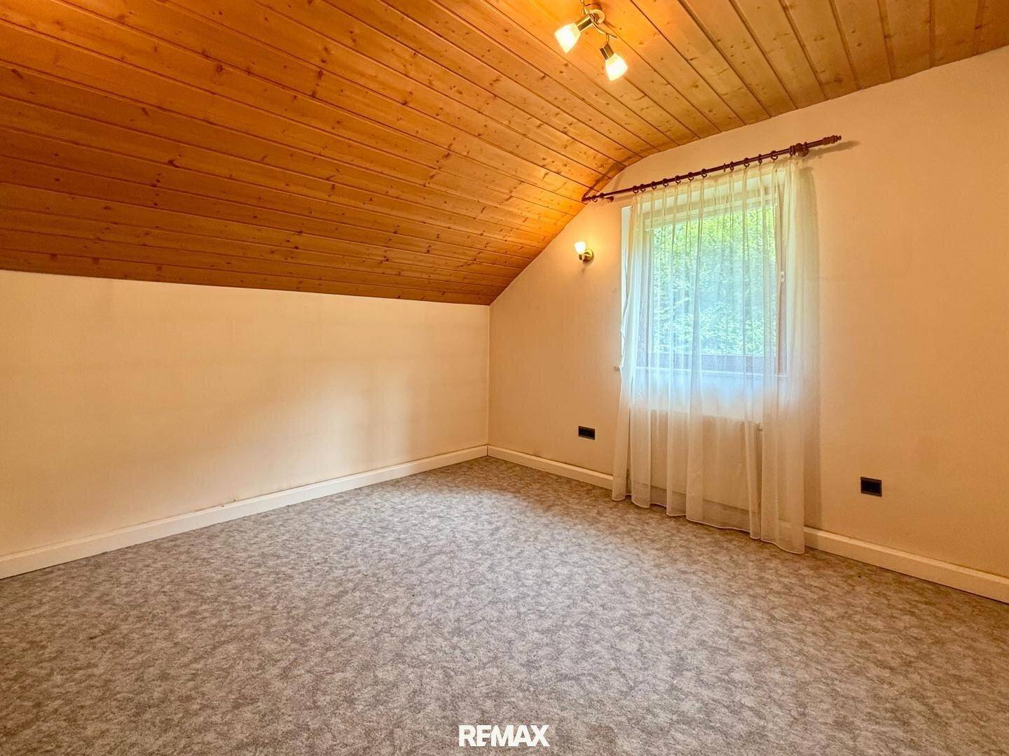Gemütliches Schlafzimmer im Dachgeschoss mit Teppichboden, Holzdecke und Fenster mit Grünblick.