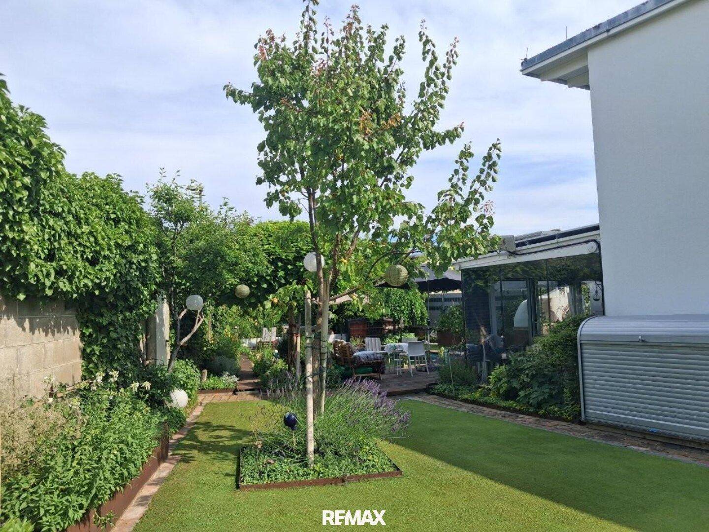 Gepflegter Garten mit Rasenfläche, Bäumen und Zugang zum verglasten Wintergarten.