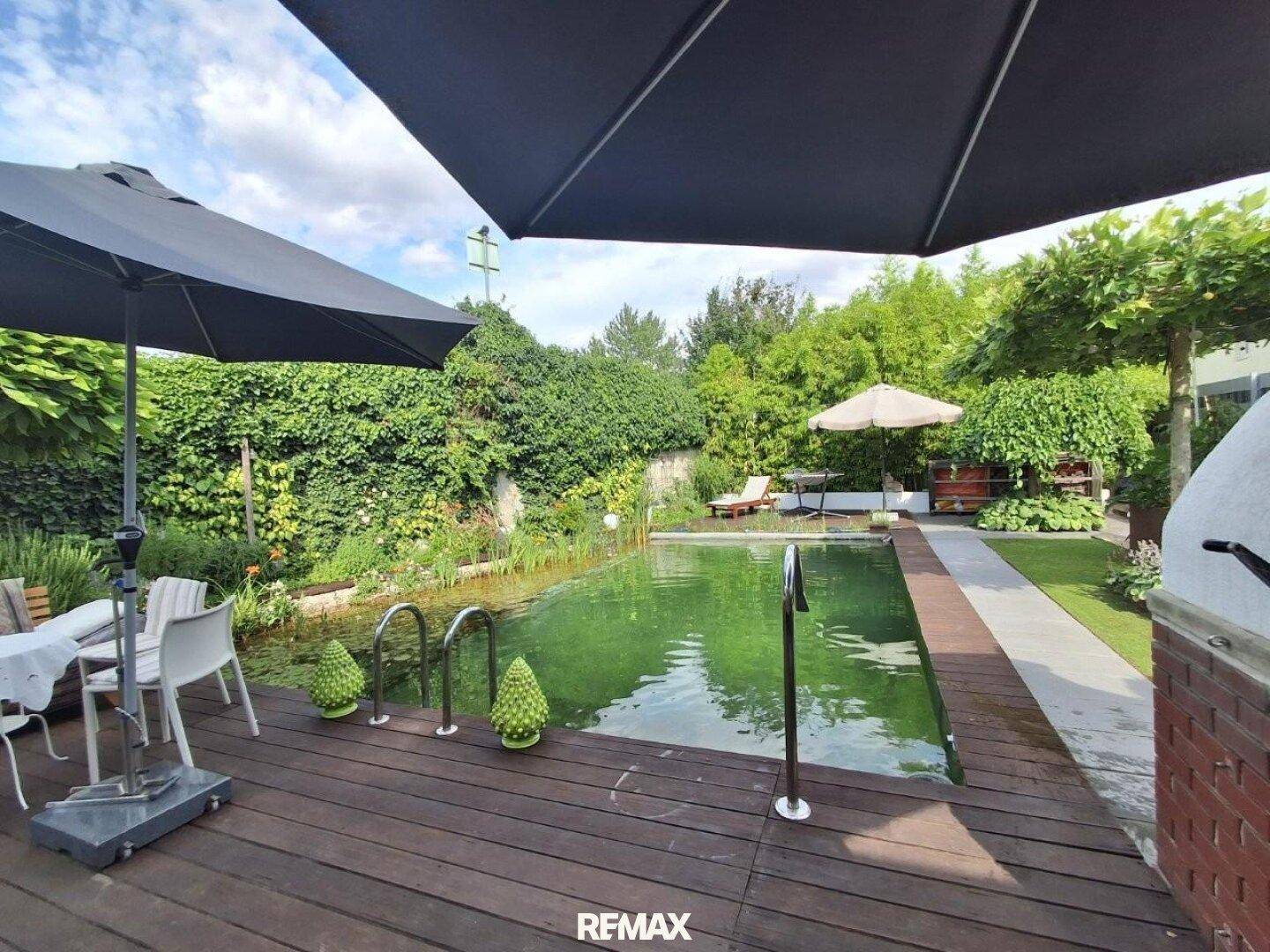 Weitläufiger Garten mit Naturpool, Holzdeck und schattigen Sitzbereichen unter Sonnenschirmen.
