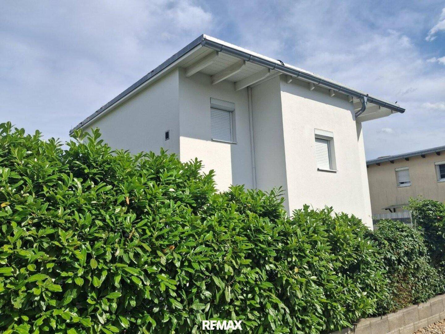 Moderne Hausfassade mit weißen Wänden und Rollläden, umgeben von einer dichten Hecke.