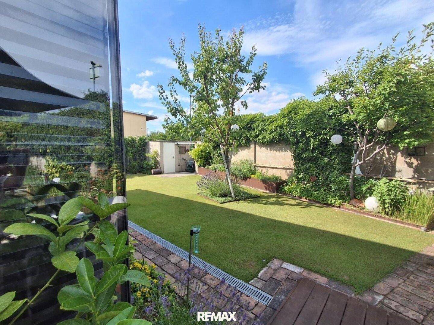 Gepflegter Garten mit Rasenfläche, Sträuchern und einem Gartenhaus unter blauem Himmel.
