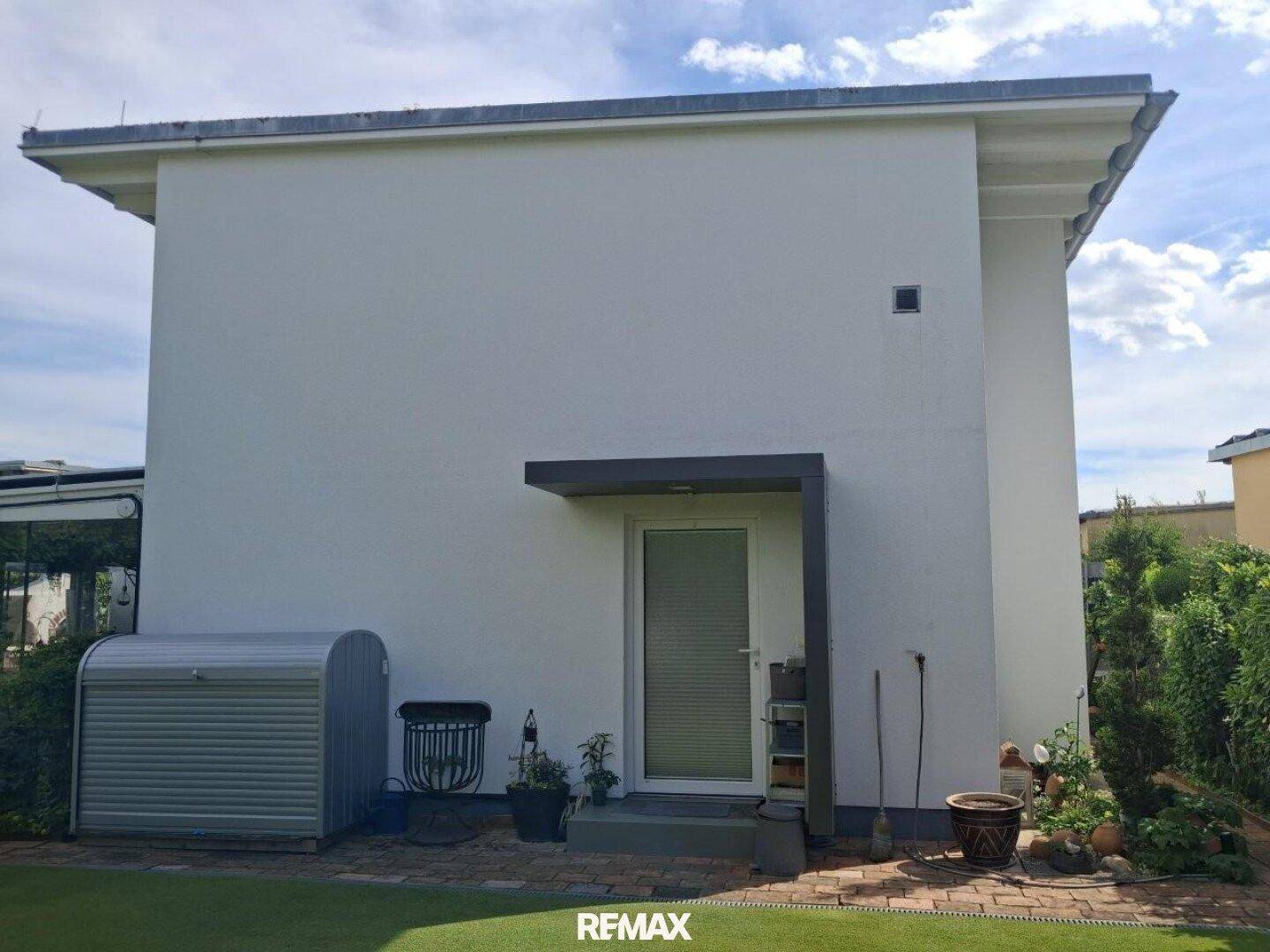 Seitenansicht des Hauses mit Zugangstür, kleinem Geräteschuppen und Rasenfläche.