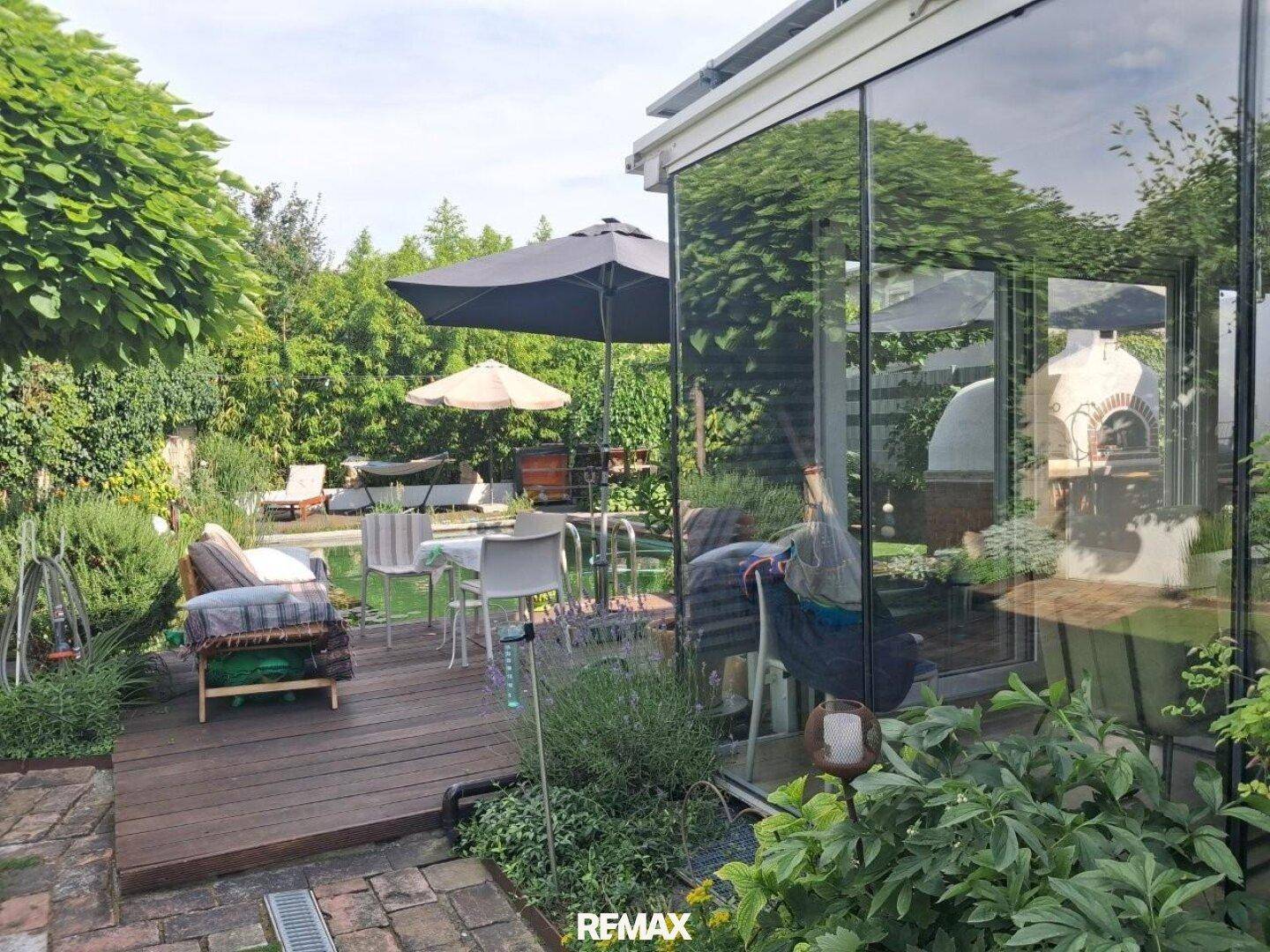 Holzterrasse mit Sitzgelegenheiten, umgeben von Grün und angrenzendem Wintergarten.