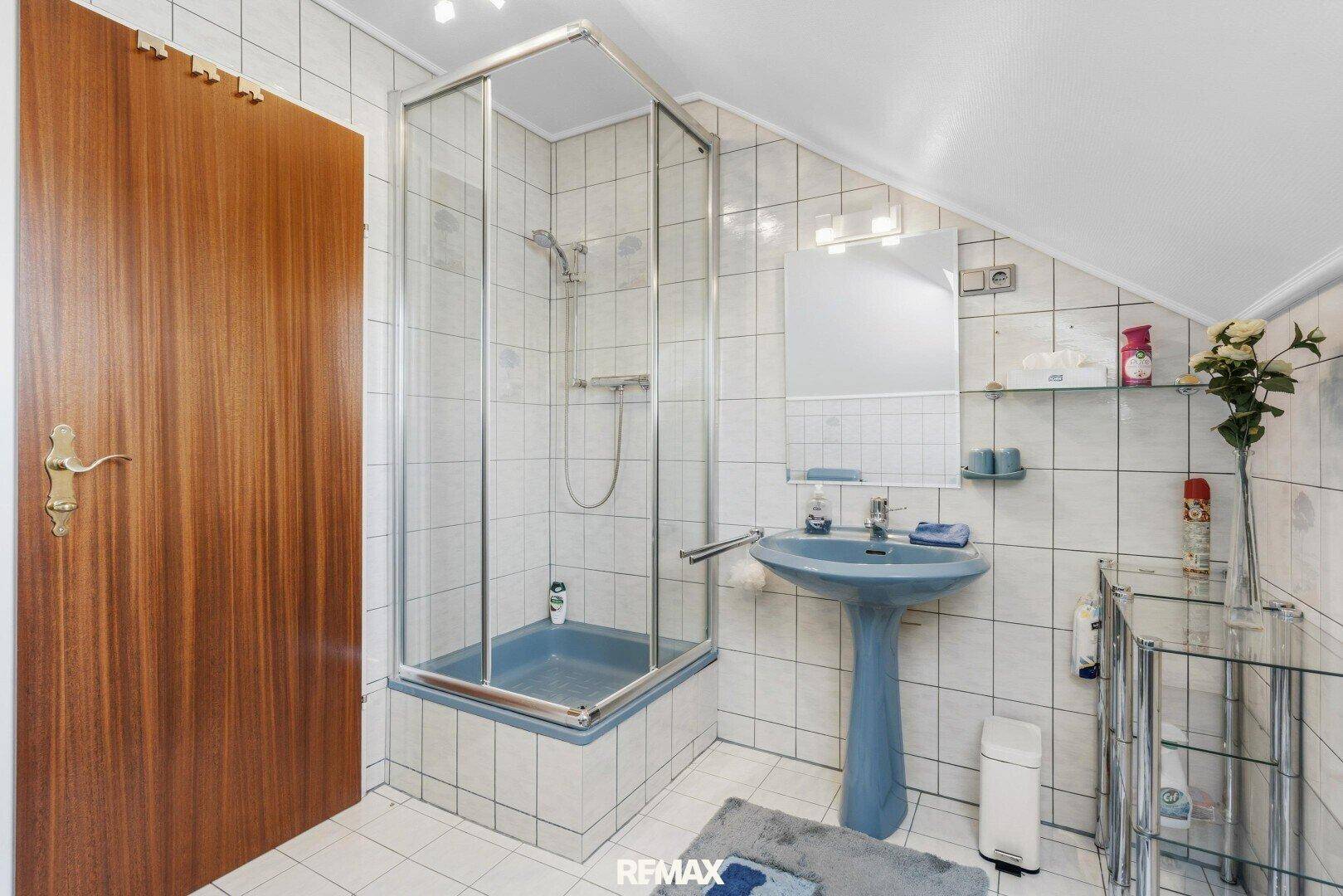 Dachgeschoss-Badezimmer mit Duschkabine, blauem Waschtisch und weißen Fliesen.