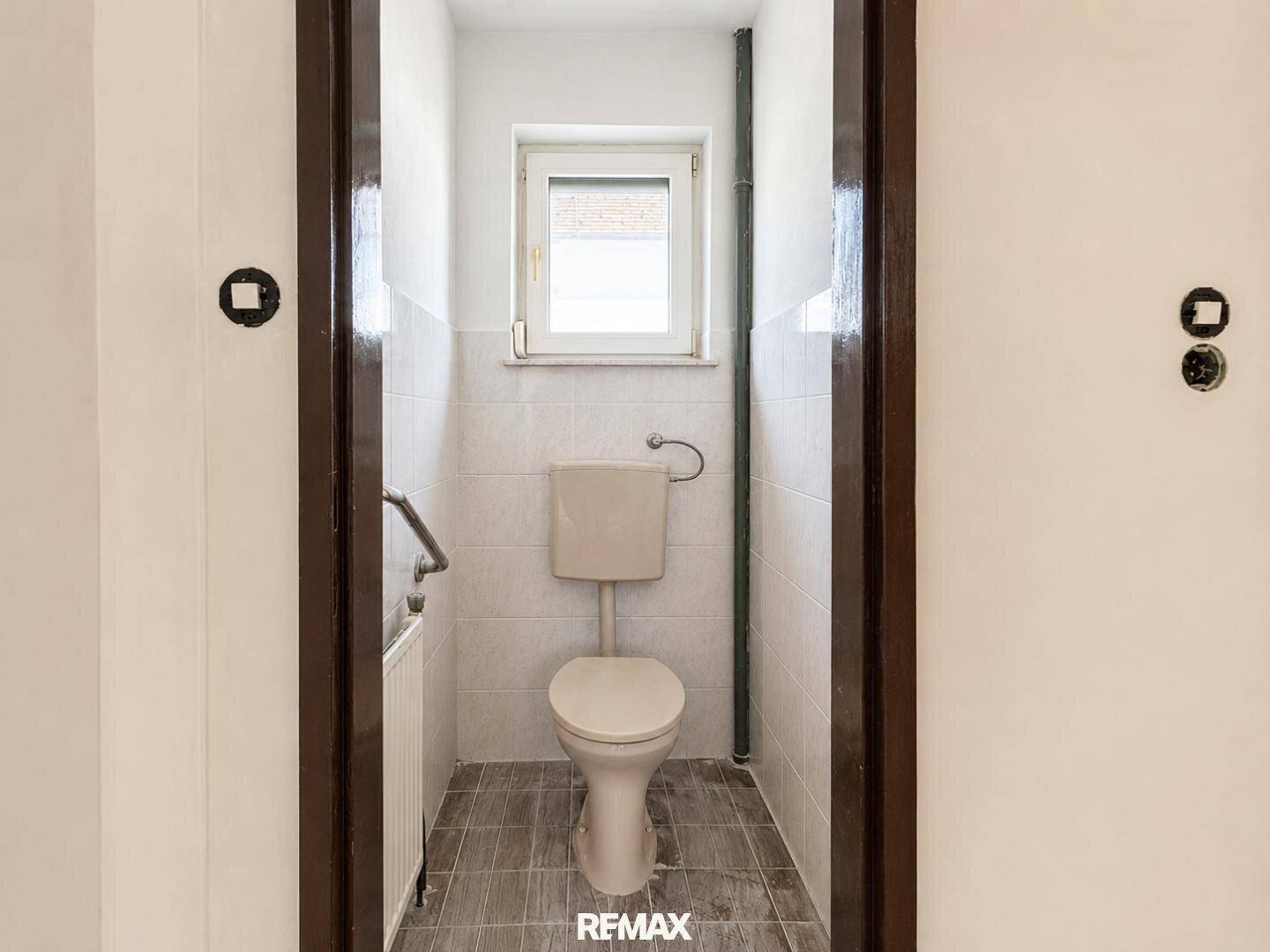 Kleines Gäste-WC mit Fenster, Toilette und hellen Fliesen an den Wänden.