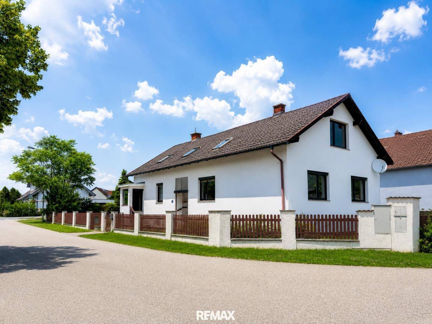 Das Haus von der Straße aus gesehen, umgeben von einem Holzzaun und viel Grün.