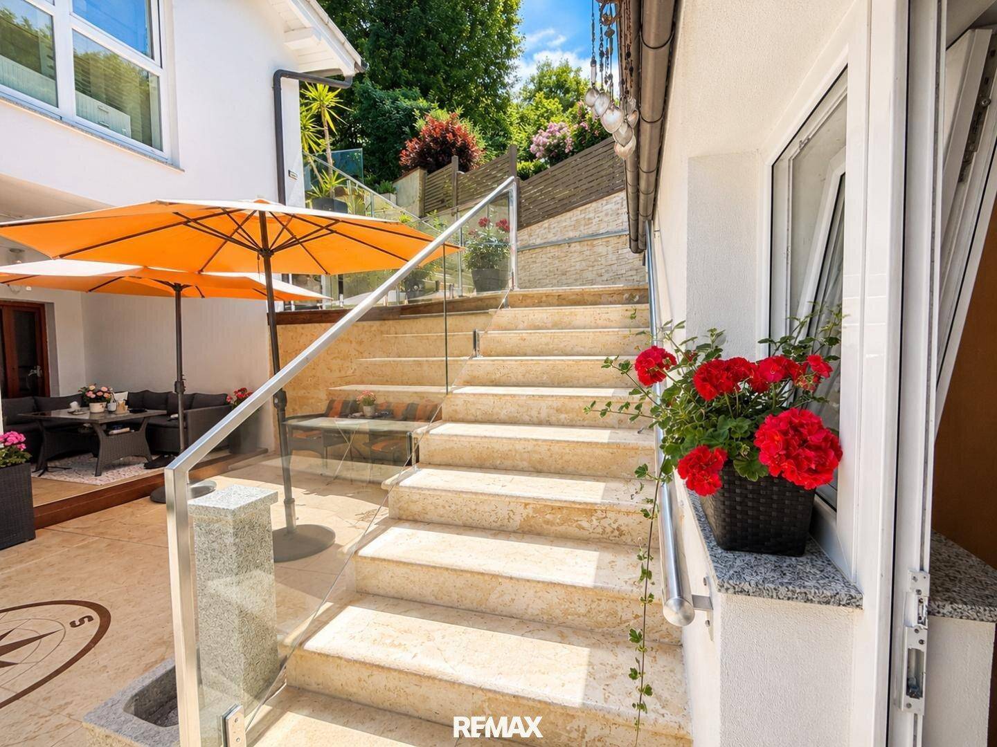 Heller Terrassenbereich mit Steintreppe, Glasgeländer und blühenden Geranien am Fenster.