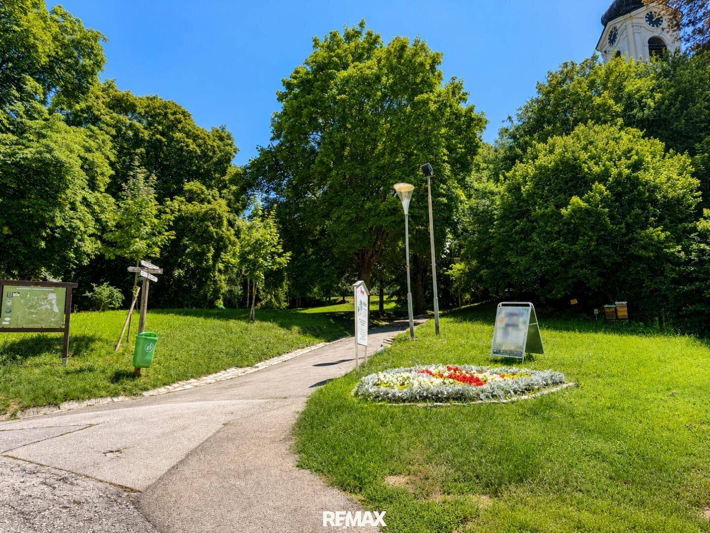 Grüne Parkanlage mit Bäumen, Rasenflächen, einem Weg und bunten Blumenbeeten.