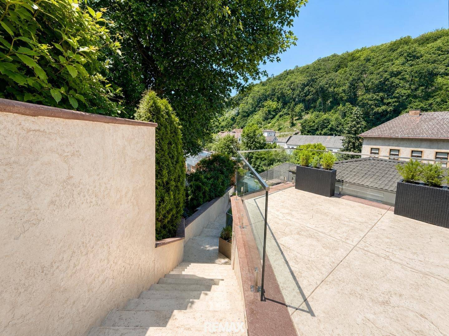 Heller Terrassenbereich mit Treppe und Glasgeländer, umgeben von üppigem Grün und Bäumen.