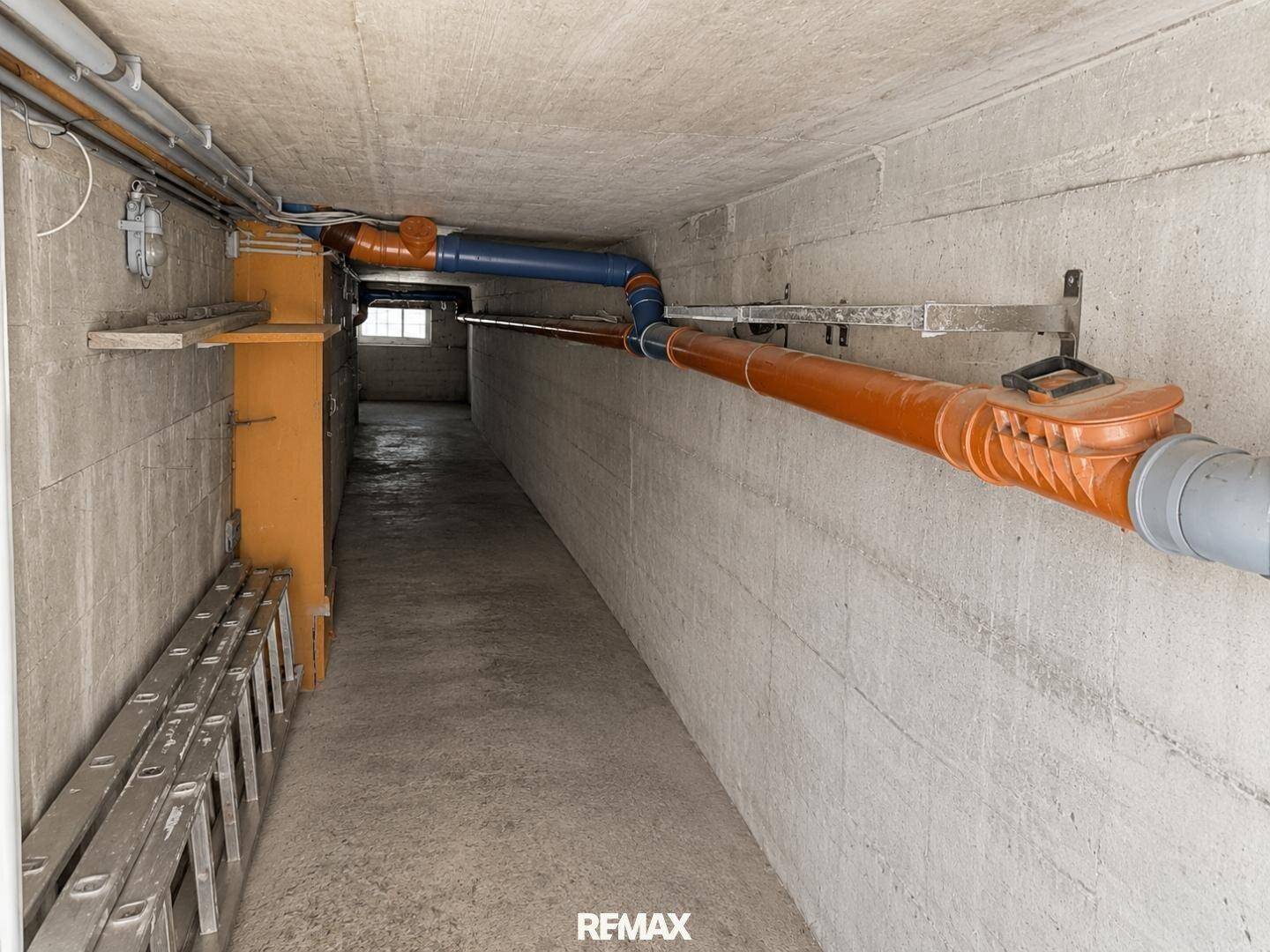 Kellerflur mit sichtbaren Rohren, Betonwänden und Lagerfläche.