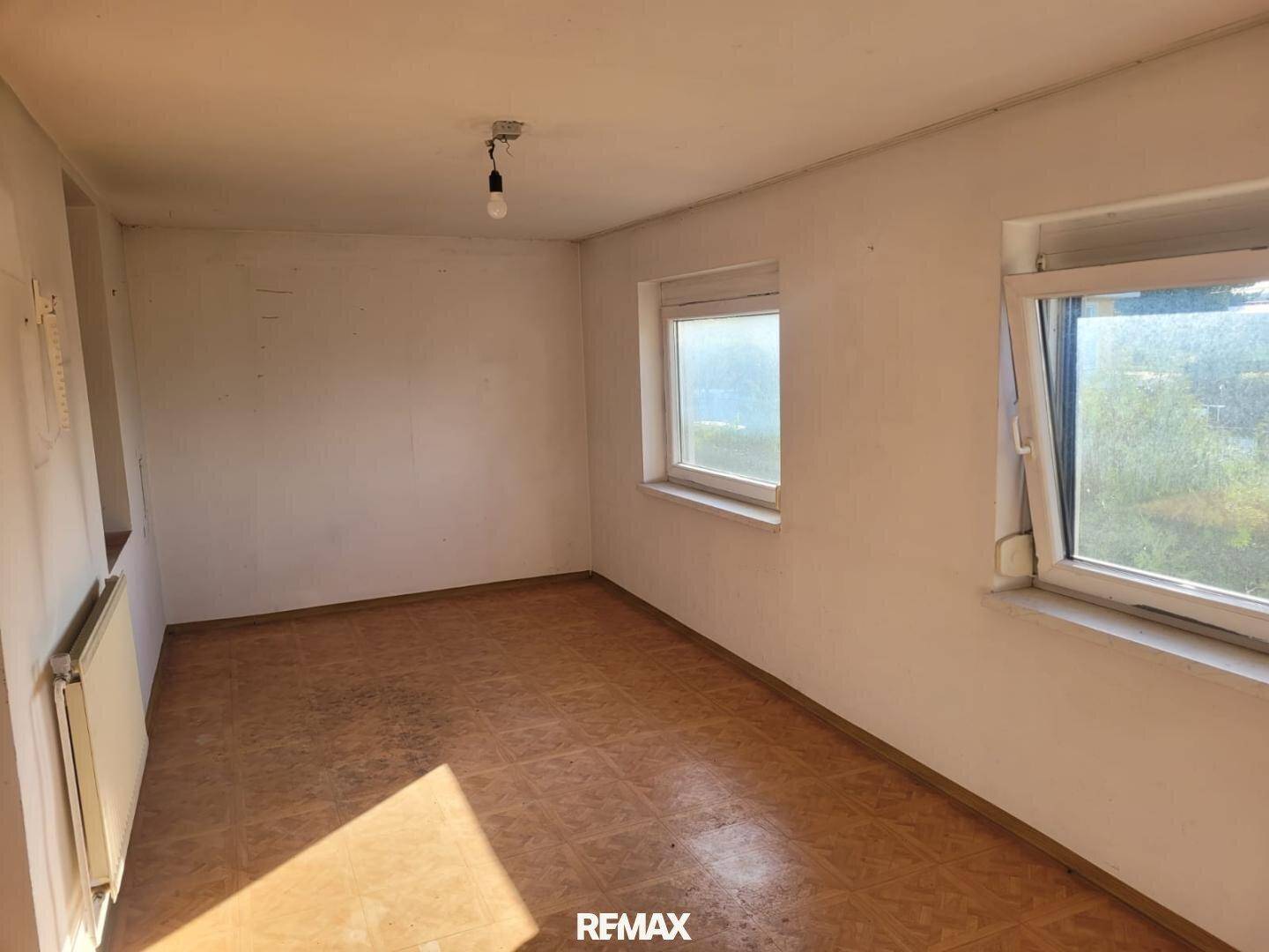 Leerer Raum mit zwei Fenstern, Heizkörper und älterem PVC-Bodenbelag, der renoviert werden muss.
