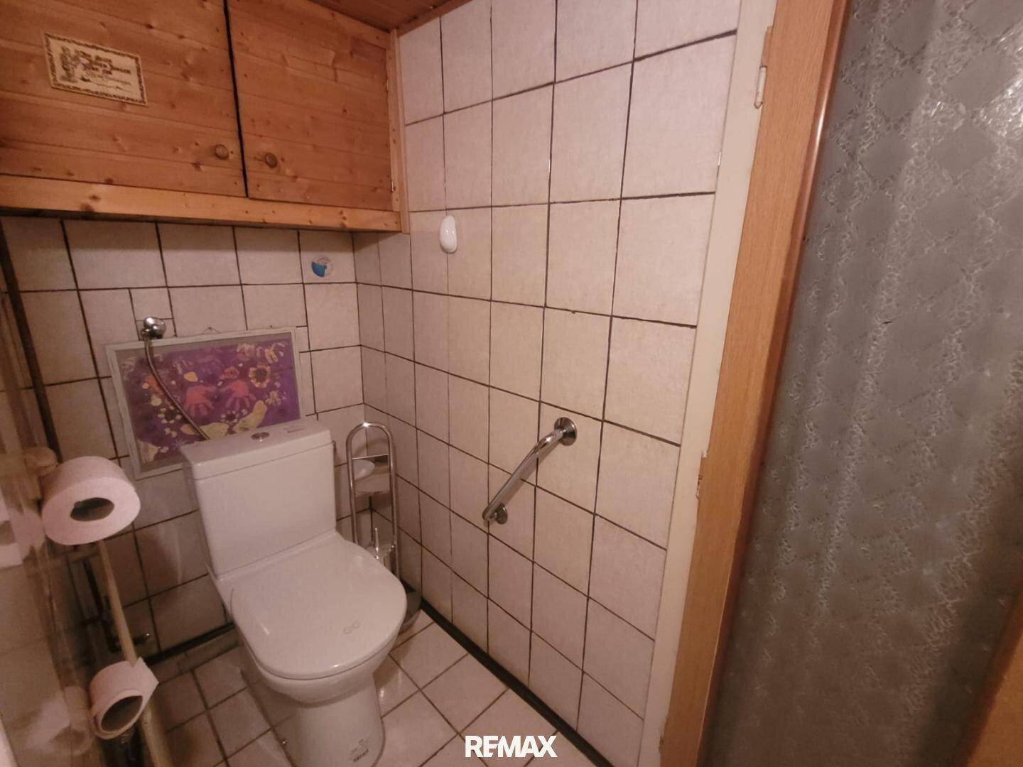 Kleines WC mit Fliesen, Toilette, Haltegriff und Holzschränken über dem Spülkasten.