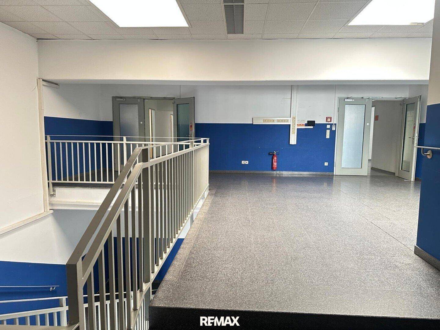 Großer Empfangsbereich mit blauen Akzentwänden, grauem Fliesenboden und Treppengeländer, hell beleuchtet.