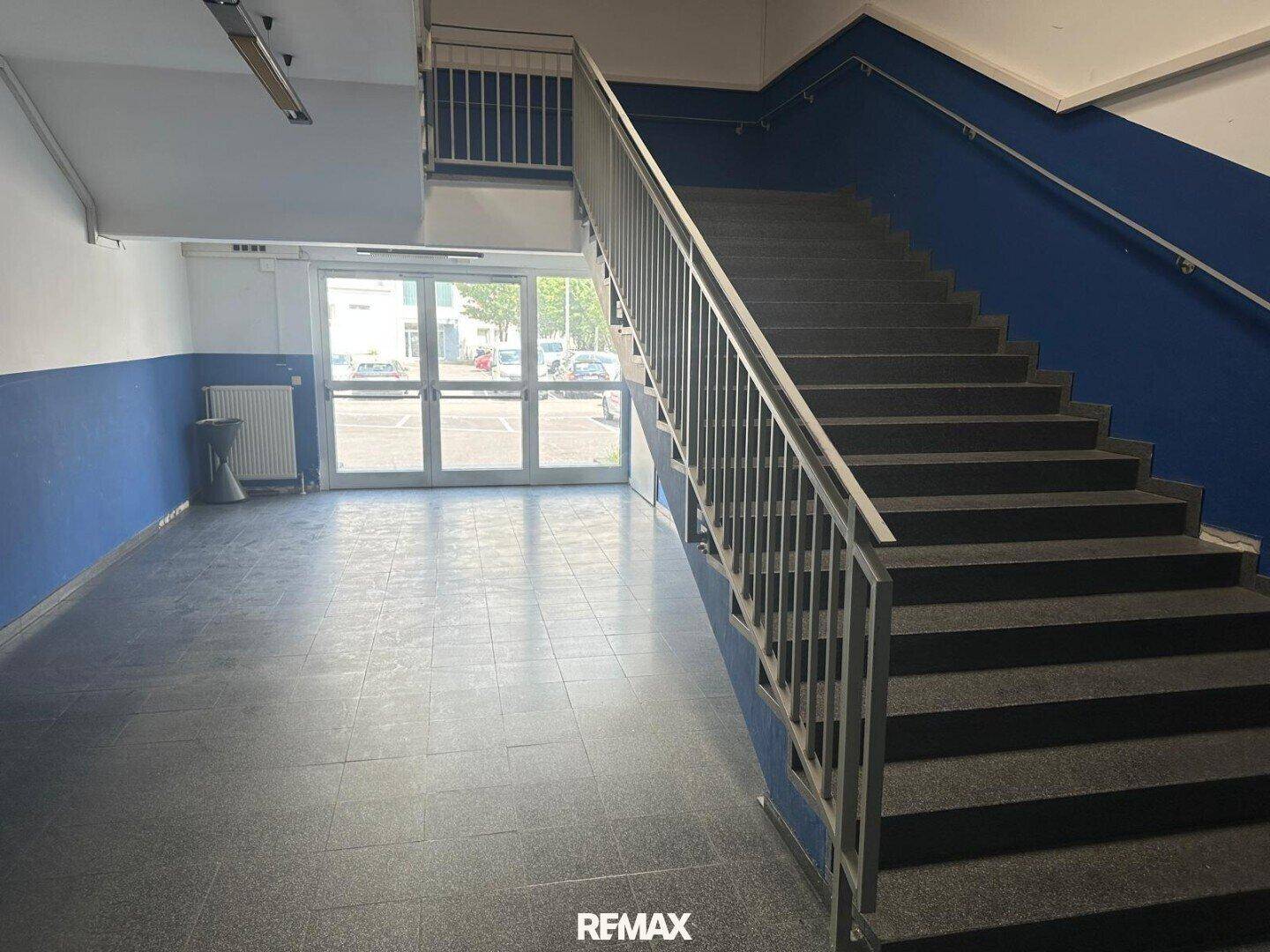 Geräumiges Treppenhaus mit grauen Stufen, blauen Wänden und einer Glastür, die zum Außenbereich führt.