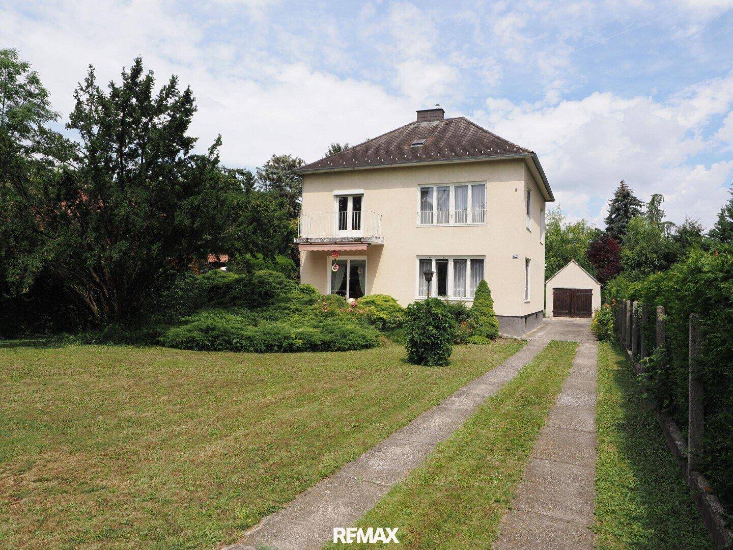 Klassisches zweistöckiges Haus mit heller Fassade, umgeben von einem großen Garten und Zufahrt zur Garage.