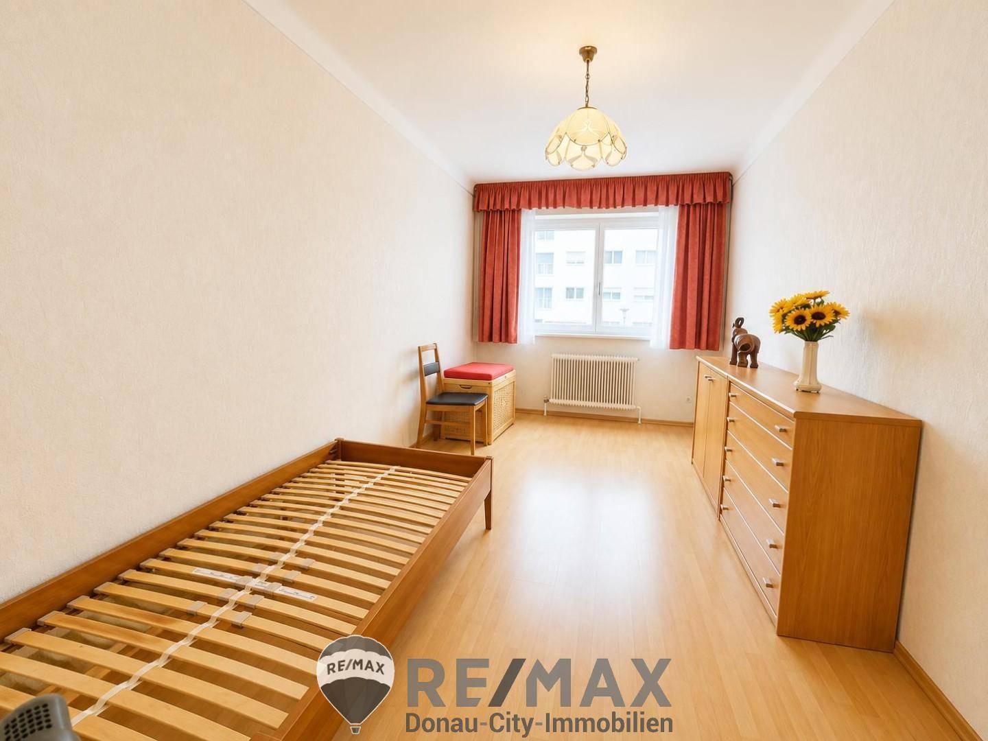 Schlafzimmer mit Parkettboden, Einzelbettrahmen und Fenster mit roten Vorhängen.