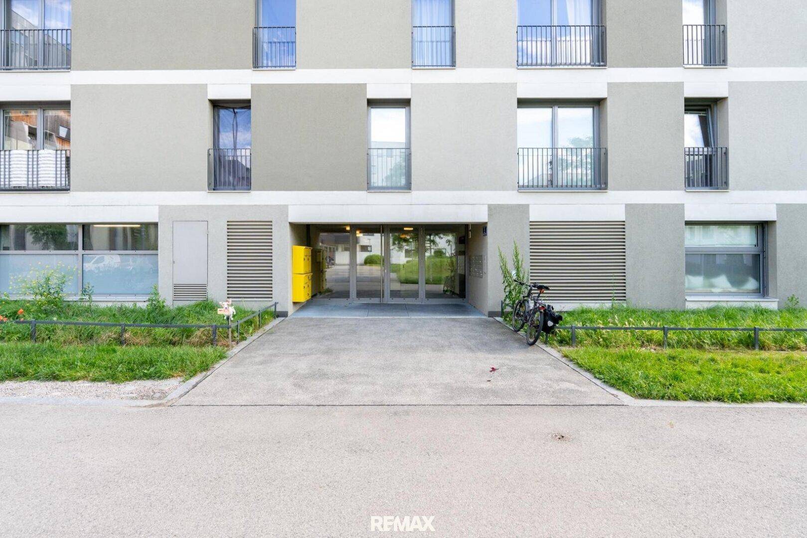 Eingangsbereich eines modernen Wohngebäudes mit Zufahrt, Fahrradabstellplatz und gepflegter Fassade.