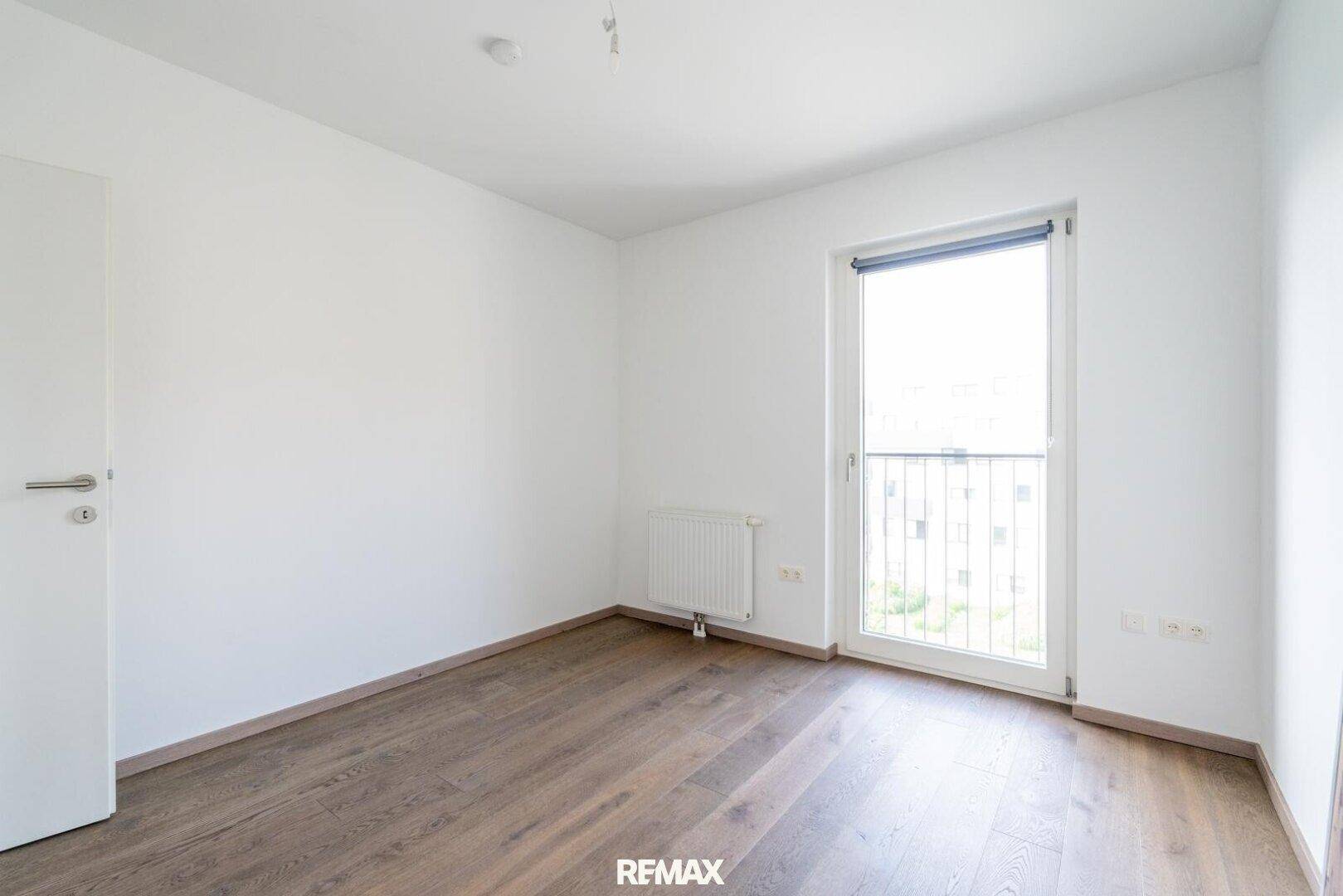 Leeres Schlafzimmer mit Holzboden, Heizkörper und Fenster mit Blick ins Grüne.