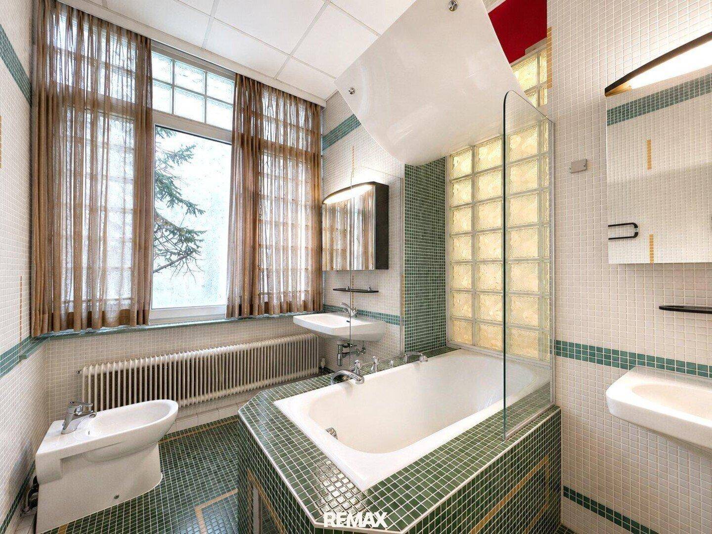 Badezimmer im 80er-Jahre-Stil mit Badewanne, Bidet, zwei Waschbecken und Fliesen in Grün- und Weißtönen.