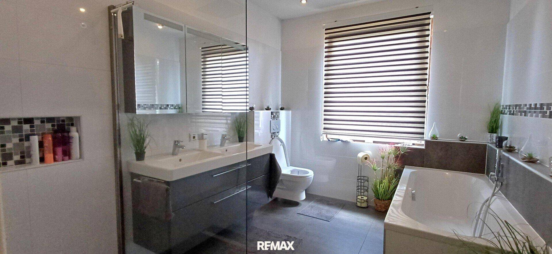 Modernes Badezimmer mit Doppelwaschbecken, Badewanne und großem Fenster. Helle Fliesen und viel Licht.