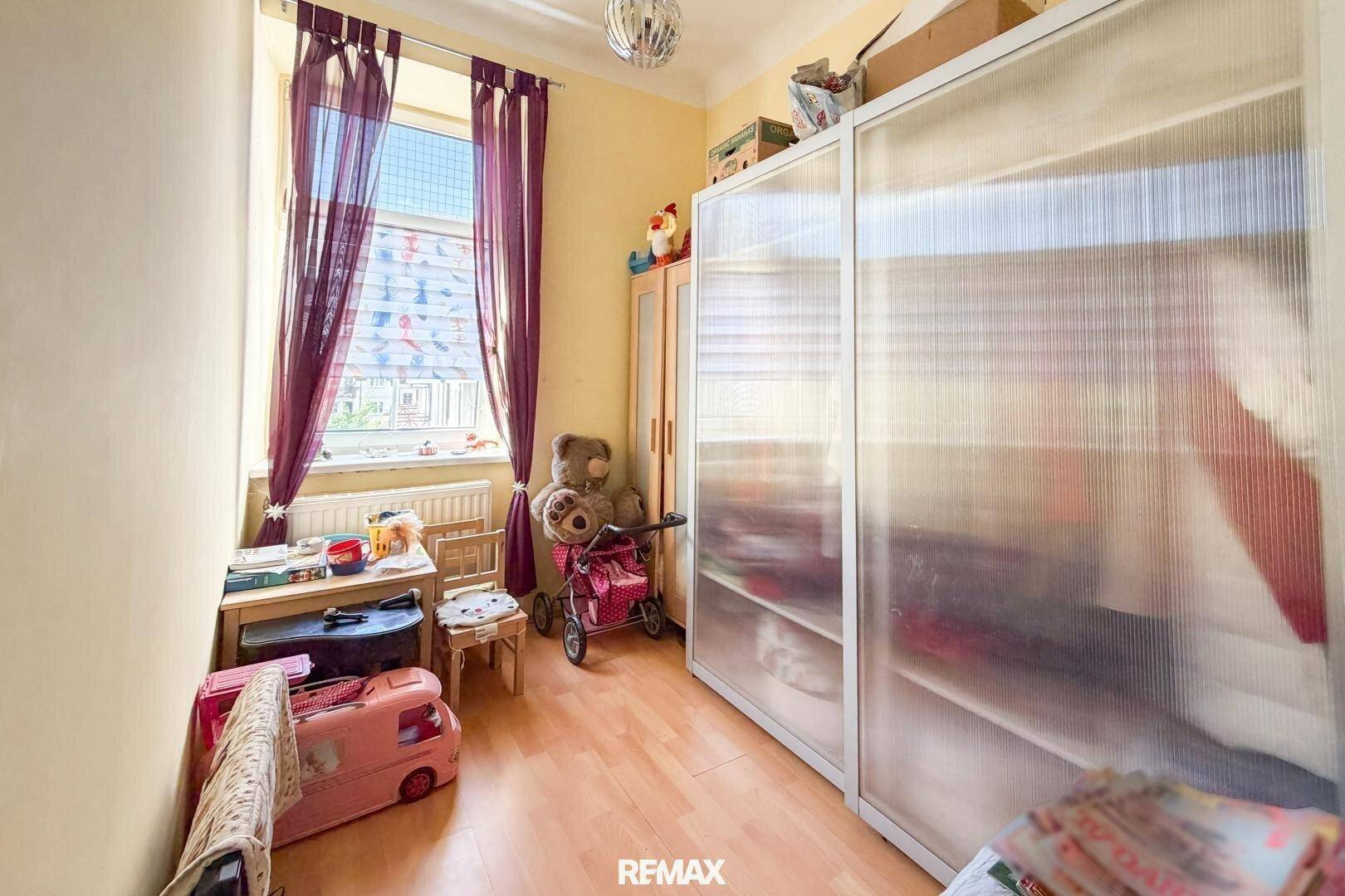 Kinderzimmer mit hellem Holzfußboden, Schrank und Fenster mit Vorhängen.