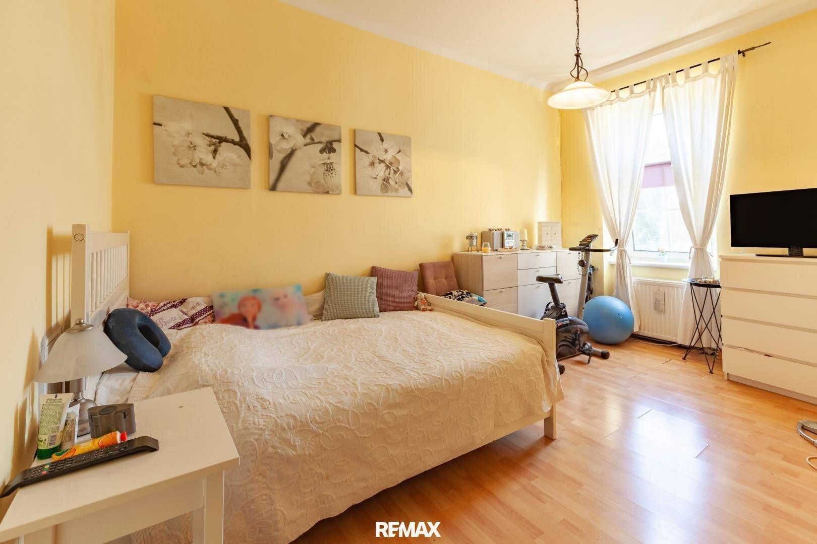 Helles Schlafzimmer mit Bett, Kommode, Trainingsgerät und Fenster mit Vorhängen.
