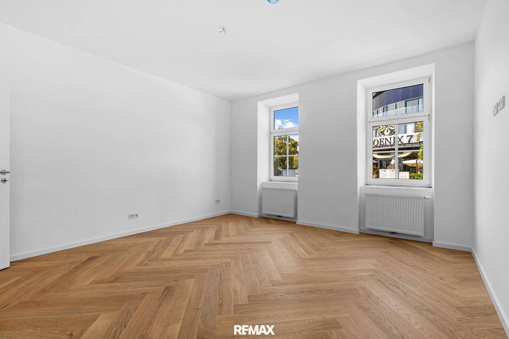 Leeres Zimmer mit Fischgrätparkett und zwei großen Fenstern, die viel Tageslicht hereinlassen.