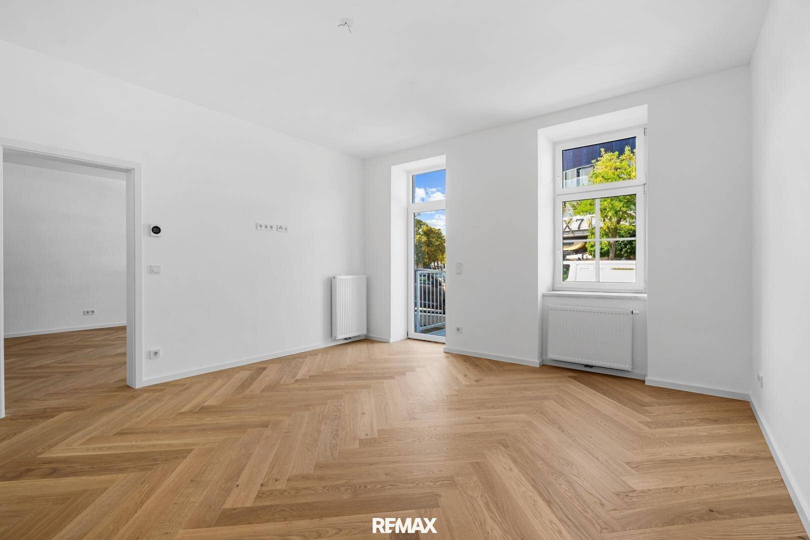 Geräumiges Wohnzimmer mit Fischgrätparkett, weißen Wänden und Zugang zu einem französischen Balkon.