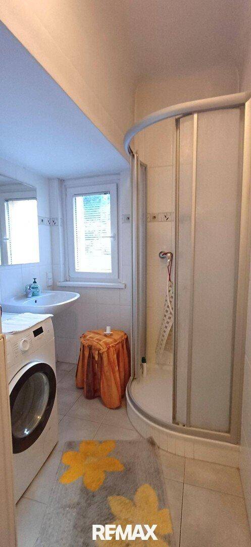 Badezimmer mit Waschmaschine, Waschbecken, Spiegel und einer Duschkabine sowie einem Fenster.