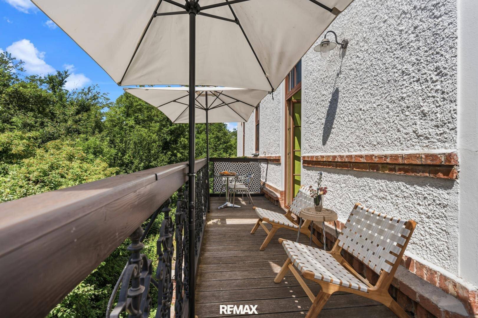 Schmaler Balkon mit Holzboden, Sitzgelegenheiten und Sonnenschirm, umgeben von Bäumen.