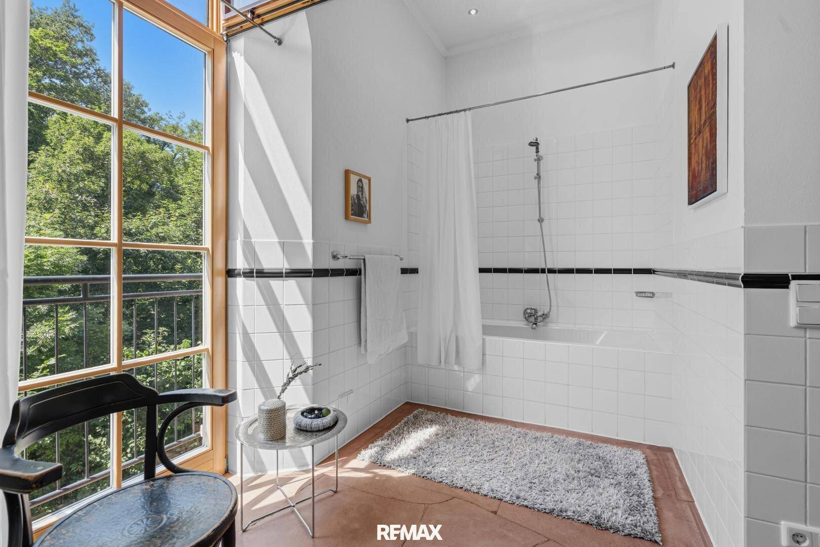 Helles Badezimmer mit Badewanne, weißen Fliesen und großem Fenster mit Blick ins Grüne.