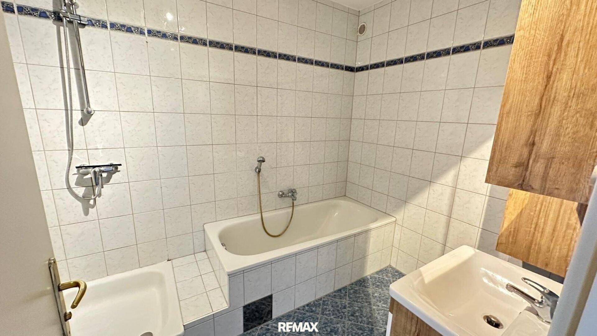 Badezimmer mit Badewanne, Duschfunktion, Waschbecken und weißen Fliesen mit blauer Bordüre.