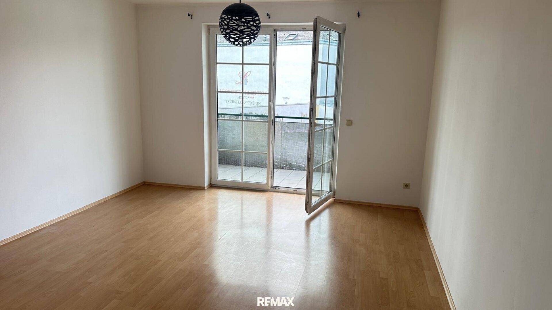 Helles Zimmer mit Parkettboden, weißen Wänden und Zugang zu einem Balkon.