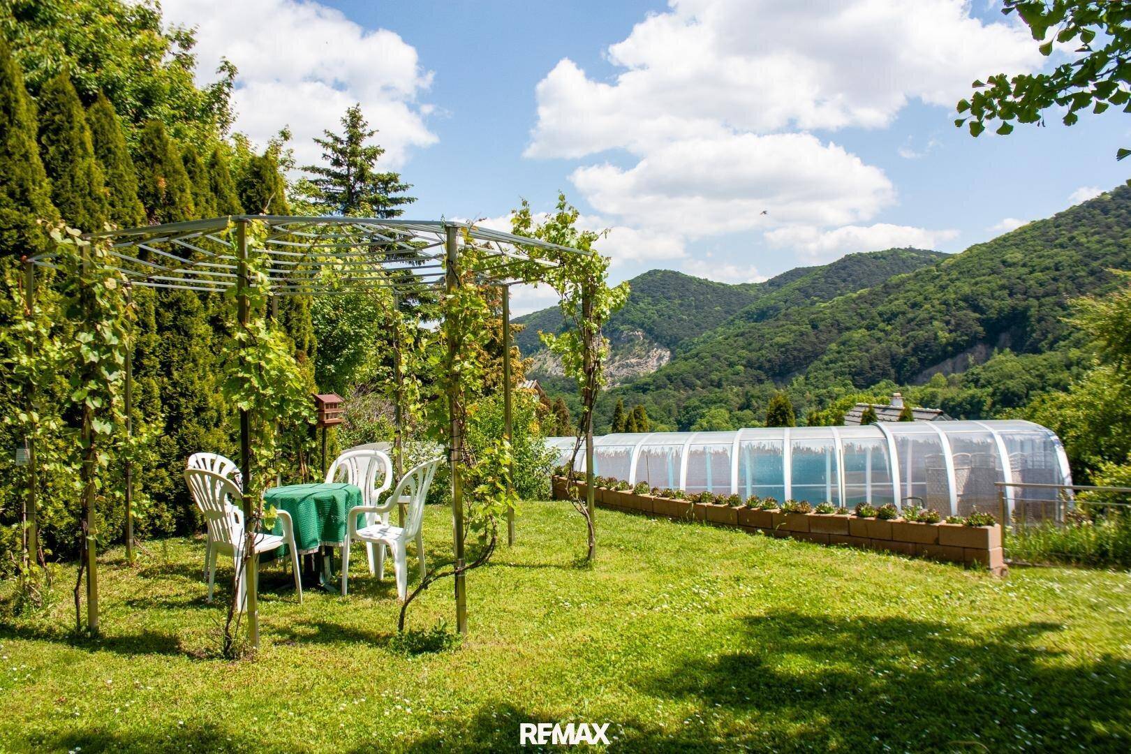 Grüner Gartenbereich mit Sitzgelegenheiten unter einer Pergola und Blick auf die bewaldeten Hügel.