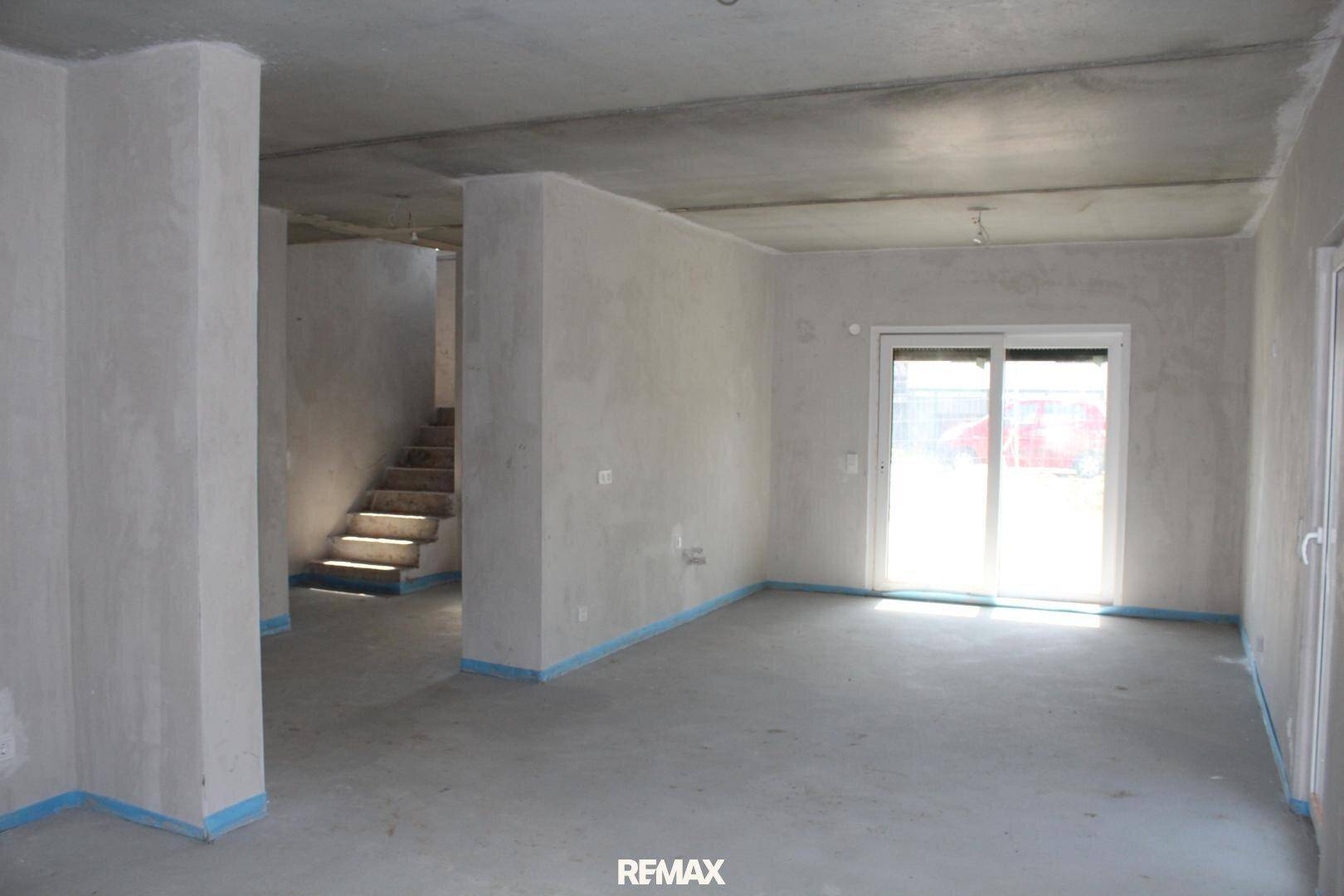 Geräumiger Rohbau-Wohnbereich mit Betonboden, Treppenaufgang und viel natürlichem Licht.
