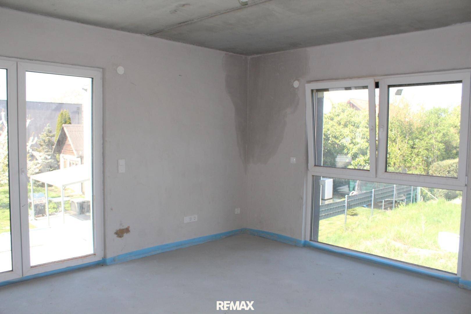 Helles Zimmer im Rohbau mit großen Fenstern und Blick ins Grüne, ideal für ein Schlafzimmer.