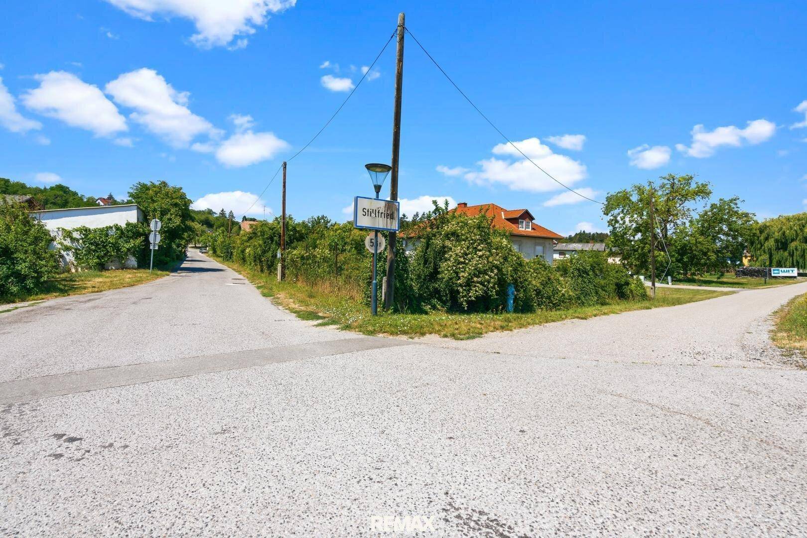 Ländliche Kreuzung mit einem Straßenschild, umgeben von Häusern und viel Grün unter blauem Himmel.