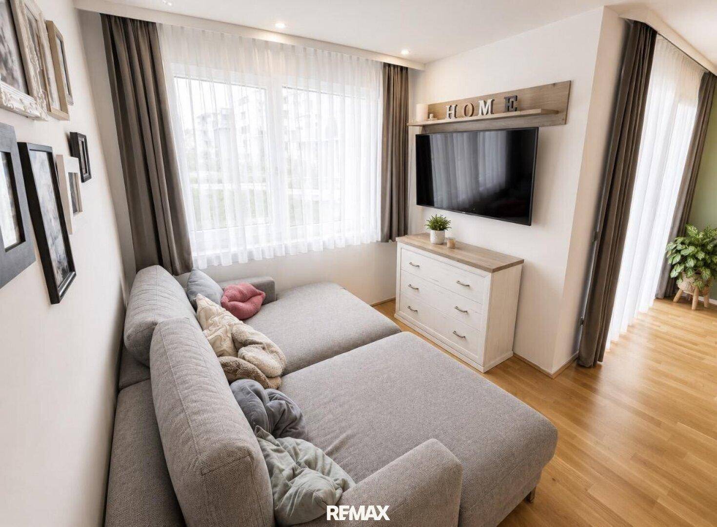 Gemütliches Wohnzimmer mit großem Sofa, Fernseher und viel Tageslicht durch das Fenster.