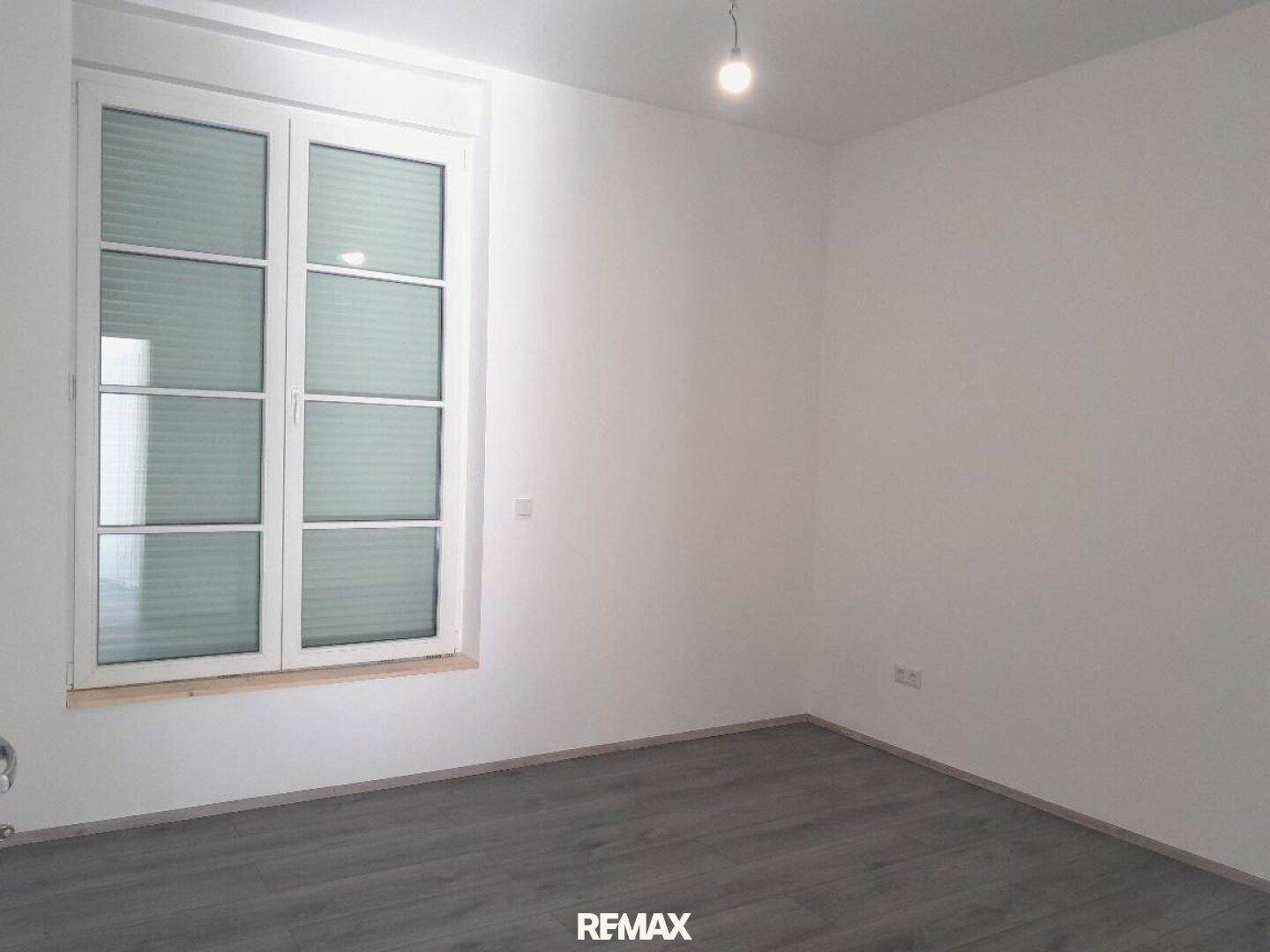 Leerer Raum mit großem Fenster, weißen Wänden und modernem grauen Bodenbelag.