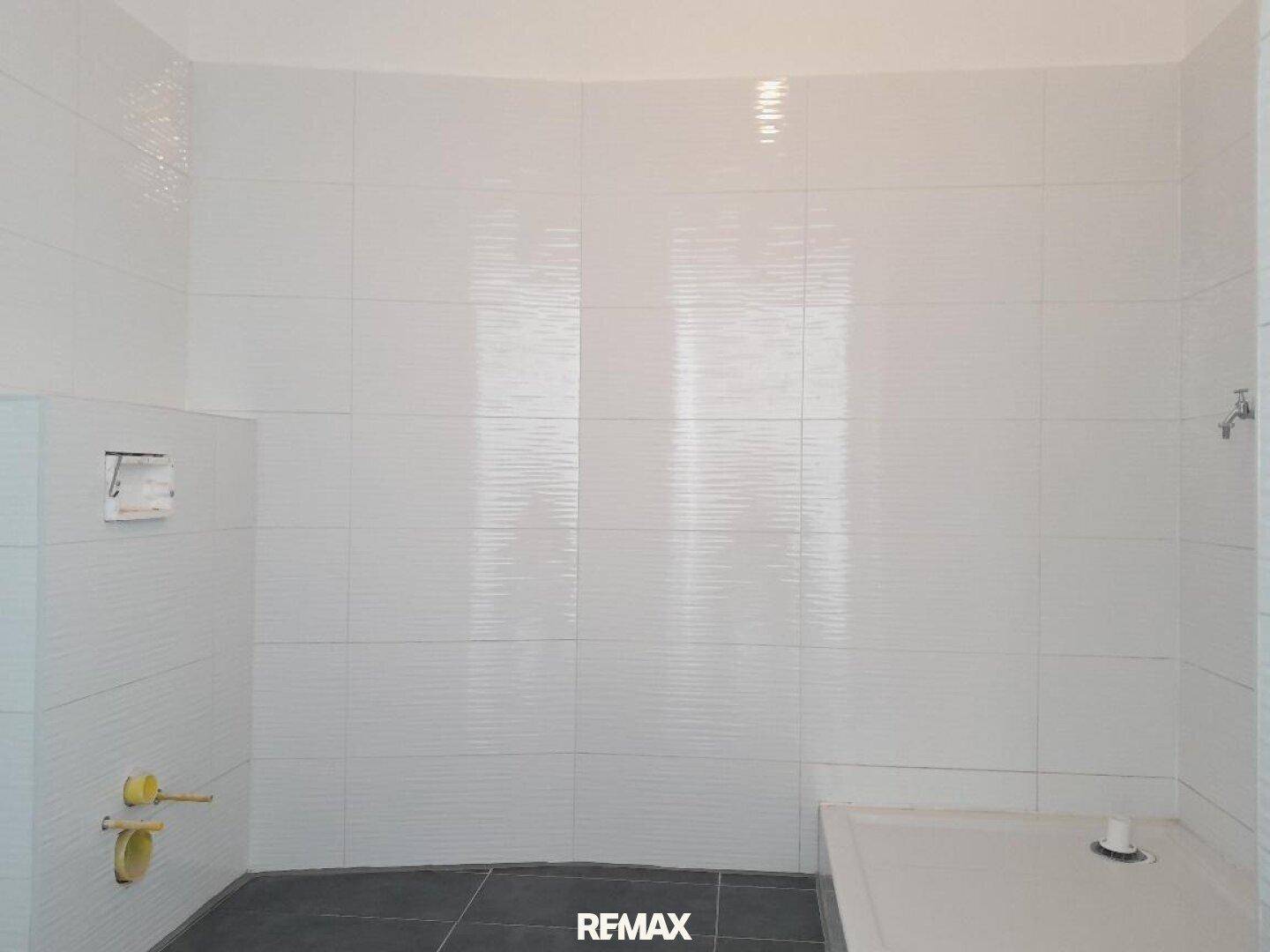 Badezimmer im Bau mit weißen Fliesen an den Wänden und dunklem Bodenbelag.