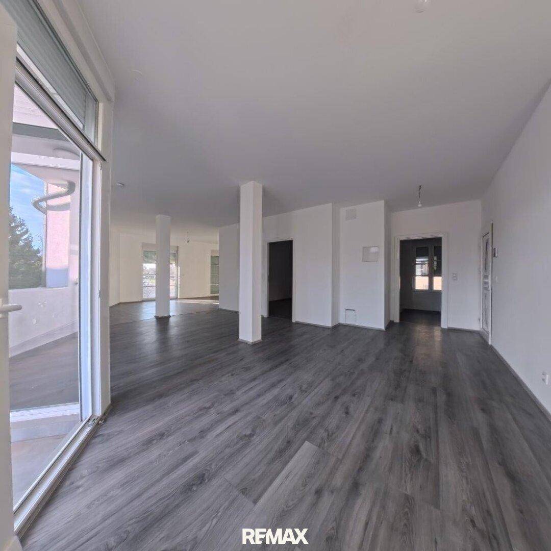 Offener, heller Wohnbereich mit Säulen, grauem Laminatboden und Zugang zu einem Balkon.