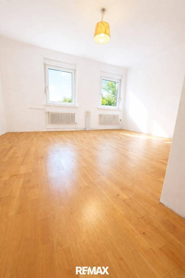 Geräumiger, leerer Raum mit Holzboden und zwei Fenstern, die viel Licht hereinlassen.
