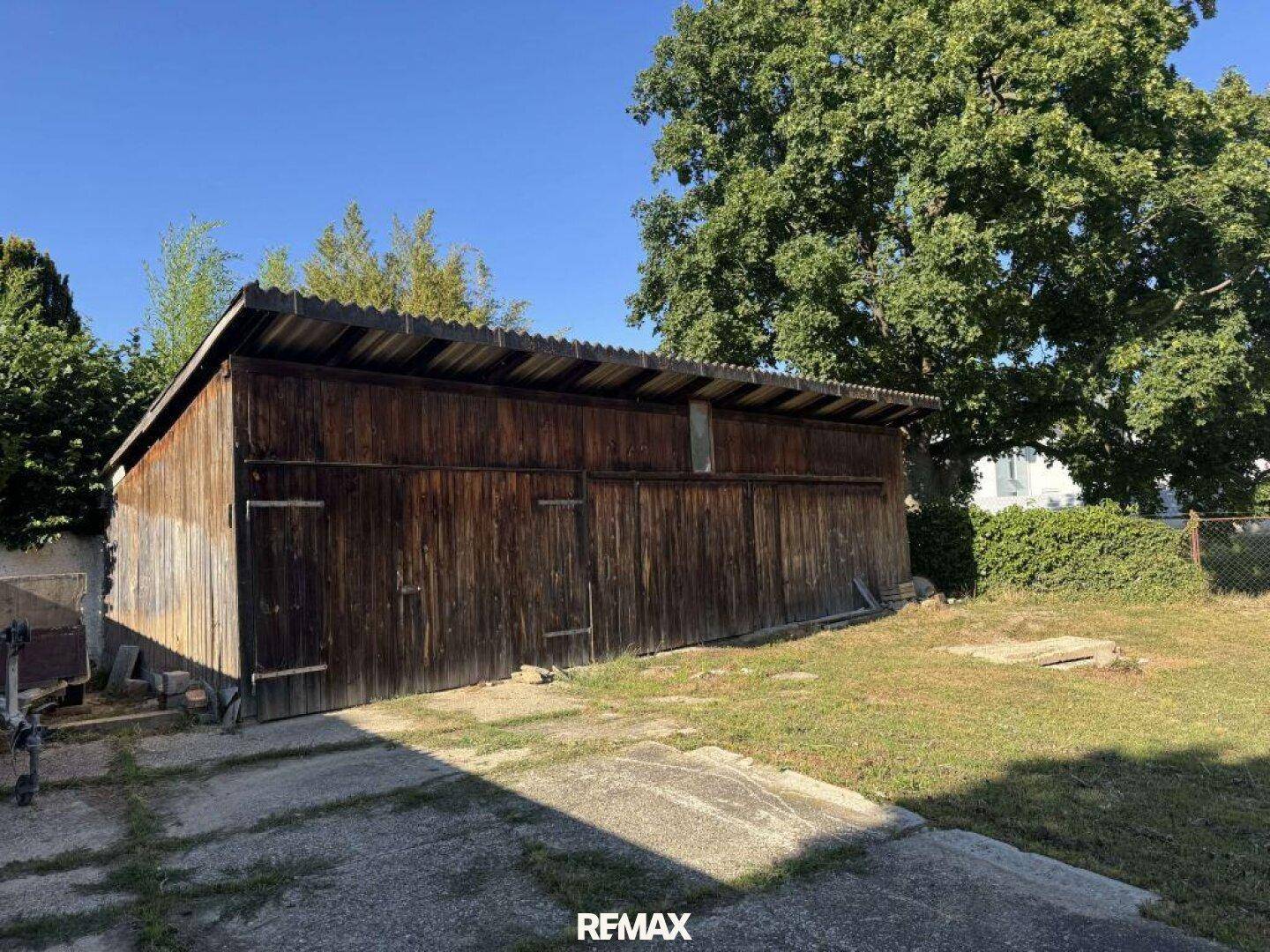 Nahaufnahme eines großen Holzschuppens mit Wellblechdach und einer betonierten Fläche davor.