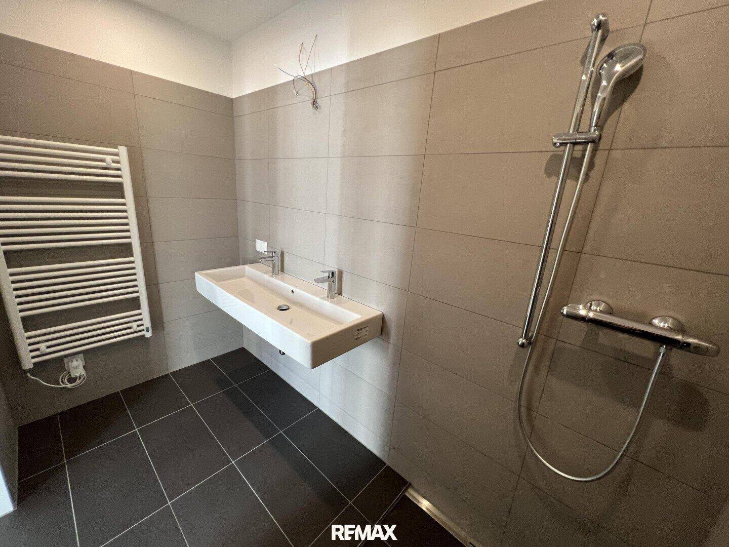Unmöbliertes Badezimmer mit Doppelwaschbecken, Duschbereich und beheiztem Handtuchhalter, modern gefliest.
