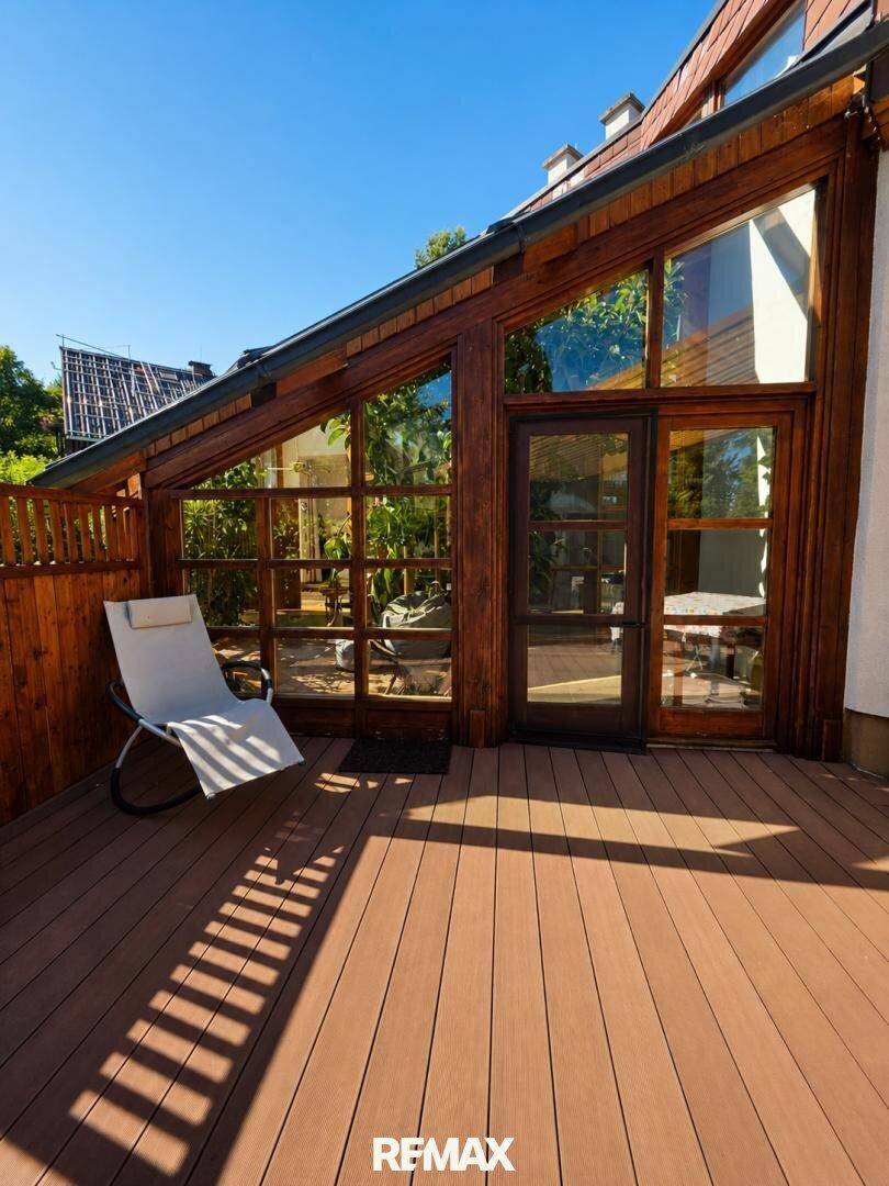 Sonnige Holzterrasse mit Liegestuhl und Zugang zum Wintergarten.