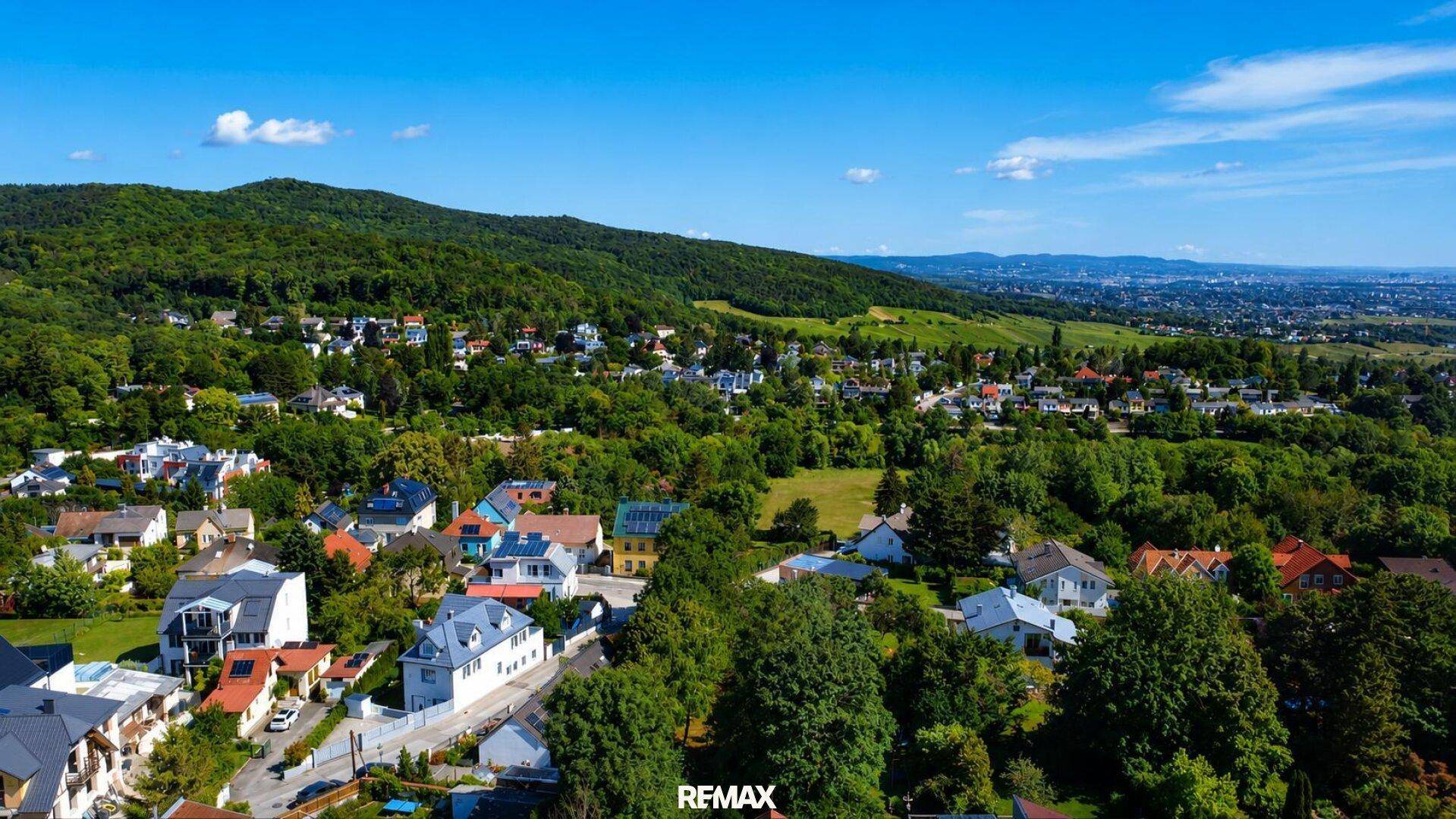 Weitläufiger Luftbildblick auf eine grüne Wohnsiedlung mit vielen Häusern und bewaldeten Hügeln.