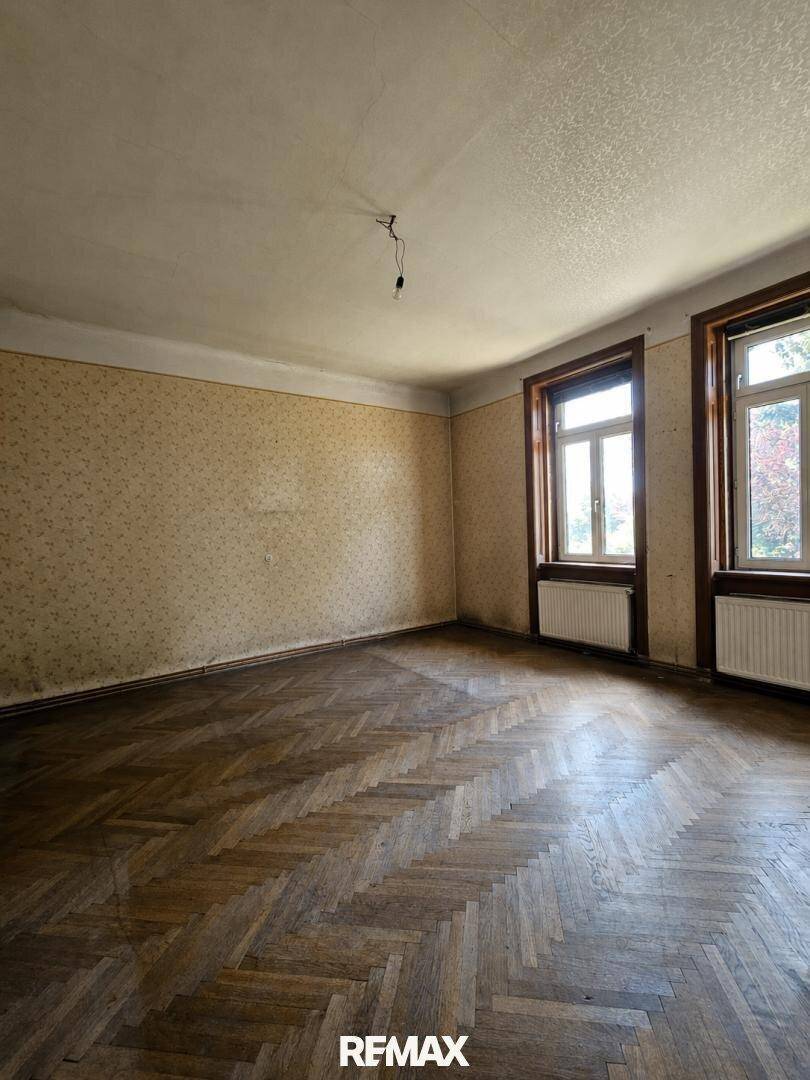Leeres Zimmer mit Fischgrätparkett, gemusterter Tapete und zwei Fenstern mit Heizkörpern darunter.