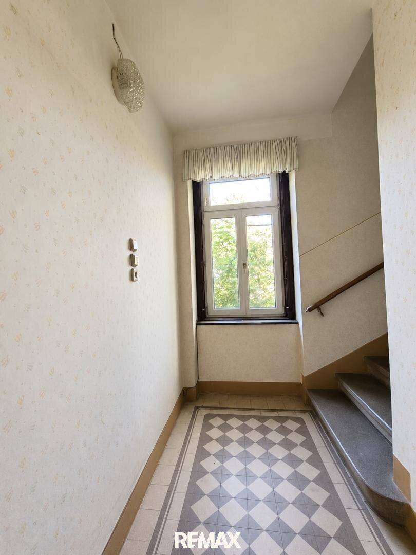 Heller Flur mit Fenster, gemusterter Tapete und Schachbrettmuster-Fliesenboden in originalem Zustand.