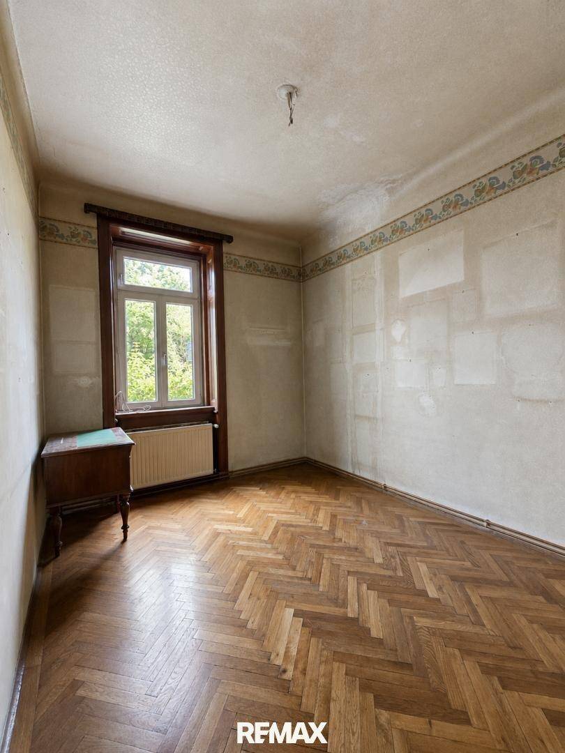 Leeres Zimmer mit Fischgrätparkett, gemusterter Tapete und einem Fenster mit Blick ins Grüne.